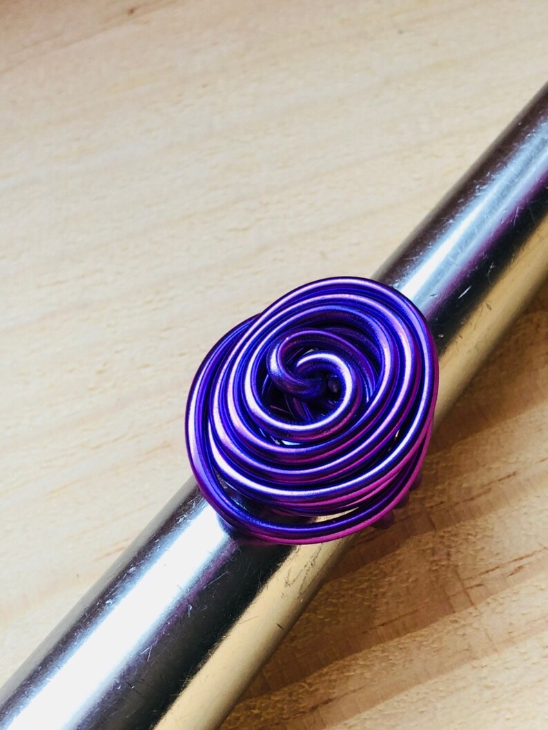K&ouml;nnte beinhalten: Ein violetter Drahtring mit einem Spiraldesign. Der Ring besteht aus mehreren Drahtwindungen, die miteinander verdreht sind, um ein einzigartiges und auff&auml;lliges Design zu schaffen.