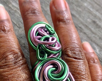 May include: A statement ring with a distinctive design of interwoven swirls. The ring is made from green and pink metallic wire, creating a striking visual effect. A unique piece of jewellery.