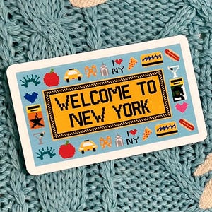 May include: A rectangular sticker with the text "WELCOME TO NEW YORK" in yellow, surrounded by icons of New York City landmarks. The sticker is set against a light blue knitted background with white accents.