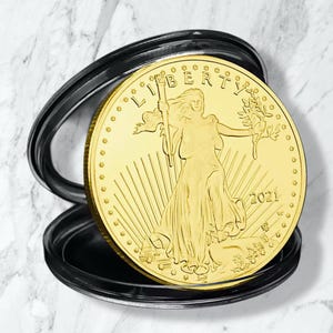 Könnte beinhalten: Eine goldene Münze mit Lady Liberty, die eine Fackel und einen Olivenzweig hält, datiert 2021. Die Münze befindet sich in einer schwarzen Schutzhülle auf einem Marmorhintergrund.