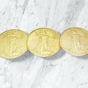 May include: Three gold-coloured coins featuring a woman holding a torch and olive branch. The coins are dated 2016, 2021, and 2023. The coins are arranged in a row on a white and grey marble surface.