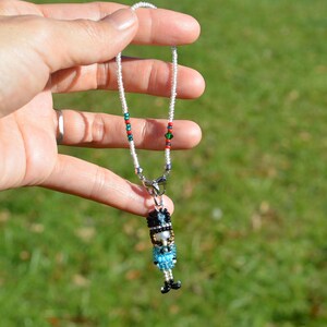 May include: A beaded necklace featuring a miniature nutcracker pendant. The necklace has a white beaded strand with red and green accents. The nutcracker pendant is detailed with black, blue, and gold beads, and black shoes.