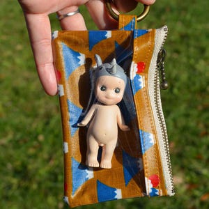 May include: A small, nude-colored figurine wearing a gray rhino helmet is partially visible inside a patterned pouch. The pouch features a brown background with blue and white geometric shapes, a zipper, and a gold-colored ring.