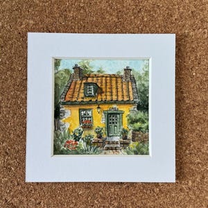 Op de afbeelding: Aquarel schilderij van een gele cottage met een oranje pannendak, groene deur en ramen. De cottage is omgeven door groen en bloemen, geplaatst in een witte passe-partout.