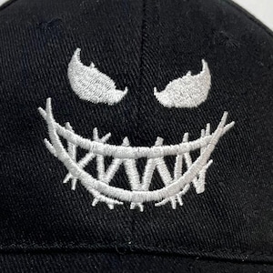 Op de afbeelding: Zwarte baseballcap met een wit geborduurd ontwerp van een dreigend gezicht. Het ontwerp heeft scherpe tanden en boze ogen. De cap heeft een gebogen klep en een gestructureerde pasvorm.