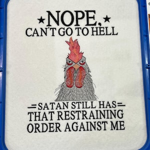 Op de afbeelding: Geborduurde witte stof met de tekst "NOPE, CAN'T GO TO HELL" en "=SATAN STILL HAS= THAT RESTRAINING ORDER AGAINST ME." Een cartoon haan met rode kam en lellen is gecentreerd onder de tekst.