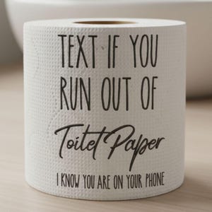 Puede incluir: Rollo de papel higiénico blanco con texto negro: "TEXT IF YOU RUN OUT OF Toilet Paper I KNOW YOU ARE ON YOUR PHONE". El rollo está sobre una superficie de madera clara.