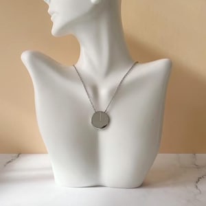 Könnte beinhalten: Silberfarbene Halskette mit rundem Anhänger und feiner Kette. Der Anhänger hat eine vertikale Linie. Die Kette wird auf einer weißen Büste vor neutralem Hintergrund präsentiert.