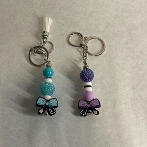 Op de afbeelding: Twee sleutelhangers met zilverkleurige hardware. Eén heeft een witte kwast, turquoise, witte en blauwe kralen en een lichtblauwe strikbedel met een stethoscoop. De andere heeft paarse, witte en lavendelkleurige kralen en een paarse strikbedel met een stethoscoop.
