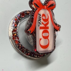 May include: A circular phone grip featuring a Diet Coke can graphic topped with a red bow. The base and the border of the graphic are filled with dark red and black glitter. The text Diet Coke is printed in red on the silver-toned soda can graphic.
