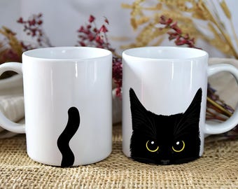 May include: Two white ceramic mugs featuring a black cat design. The left mug displays a black cat tail, while the right mug shows the face of a black cat with large yellow eyes. Both mugs have a smooth finish and a standard handle on the side.