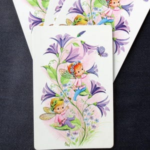 May include: Set of four vintage playing cards featuring illustrations of fairies among purple flowers and butterflies. The cards have a white background with a floral design and a small fairy illustration. The cards are rectangular and have rounded corners.