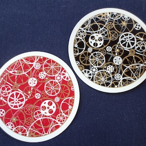 May include: Two round white coasters with a gold and white gear pattern. One coaster has a red background and the other has a black background.