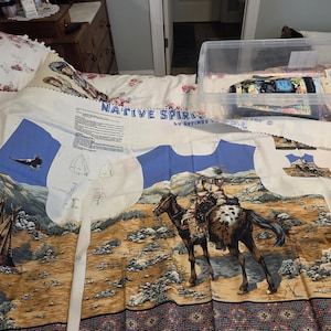 May include: A white fabric panel titled Native Spirit by Springs Creative featuring printed patterns for sewing. The design includes a person riding a spotted horse, a wolf, teepees, and a flying eagle in a desert landscape with blue sky accents.