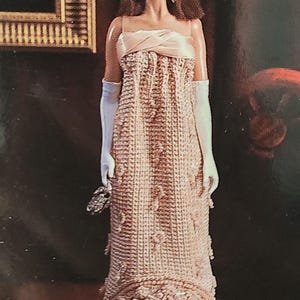 Könnte beinhalten: Eine Puppe trägt ein langes, weißes, gehäkeltes Kleid mit einem Satin-Oberteil. Das Kleid hat einen Perlenbesatz und die Puppe trägt weiße Handschuhe.