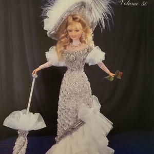 Puede incluir: Una muñeca con un disfraz blanco de ganchillo con sombrero y sombrilla a juego. La muñeca lleva un vestido largo y adornado con una falda en capas. El sombrero está adornado con plumas y detalles plateados. El texto "Crochet Collector Costume Volume 50" es visible.