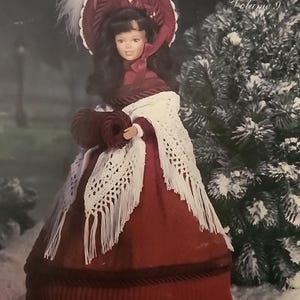 May include: A doll in a burgundy crochet costume, including a dress, bonnet, muff, boots, and fringed shawl. The doll is wearing a white shawl and a burgundy hat with white feathers. The text "Crochet Collection Costume Volume 9" is visible.