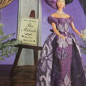Pode incluir: Uma boneca de crochê vestida com um vestido roxo com uma sobreposição de renda branca e um chapéu combinando. A boneca está segurando uma pequena bolsa de contas. A boneca está em pé em frente a uma placa que diz "Now Showing The Mikado Balcony Mezzanine".