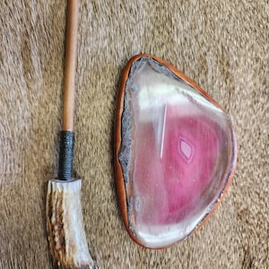 May include: A handcrafted pipe with an antler stem and a wooden mouthpiece, next to a pink and white agate stone with a leather border. The agate has a polished surface and a natural, organic shape.