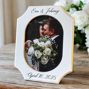 May include: White picture frame with gold trim, holding a wedding photo. The frame is inscribed with "Erin & Johnny" at the top and "April 16, 2025" at the bottom. The photo shows a couple and a bouquet of white roses.