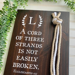 May include: A dark stained wooden sign with white text that reads "A CORD OF THREE STRANDS IS NOT EASILY BROKEN." The sign includes the text "Ecclesiastes 4:12" and the names "Jon & Lindsey" with the date "07/09/2022". Decorative rope hangs from the top.