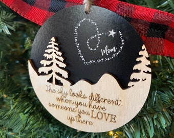 May include: A wooden Christmas ornament with a black background and a white heart with the word "Mom" inside. The ornament has a white tree and the text "The sky looks different when you have someone you LOVE up there".