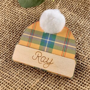 May include: A personalized Christmas ornament shaped like a hat with a plaid pattern in shades of yellow, green, blue, and red. The hat has a white pom-pom on top and a wooden tag with the name "Ray" engraved on it.