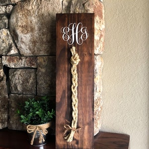 May include: A dark stained wooden sign with a decorative rope design. The sign features a white monogram and a long, braided rope element. The rope is tied at the top and bottom, creating a nautical or rustic aesthetic.