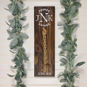 May include: A wooden sign with a brown stain and white lettering. The sign has a monogram of the letters "JNK" with a wreath of leaves around the letters. The date "2/20/2020" is written below the monogram. A brown and beige rope is attached to the sign.