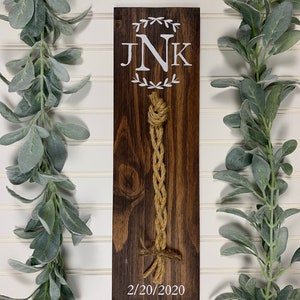 May include: A wooden sign with the initials "JNK" and a date, "2/20/2020", in white paint. A brown rope is tied in a knot and hangs from the sign.