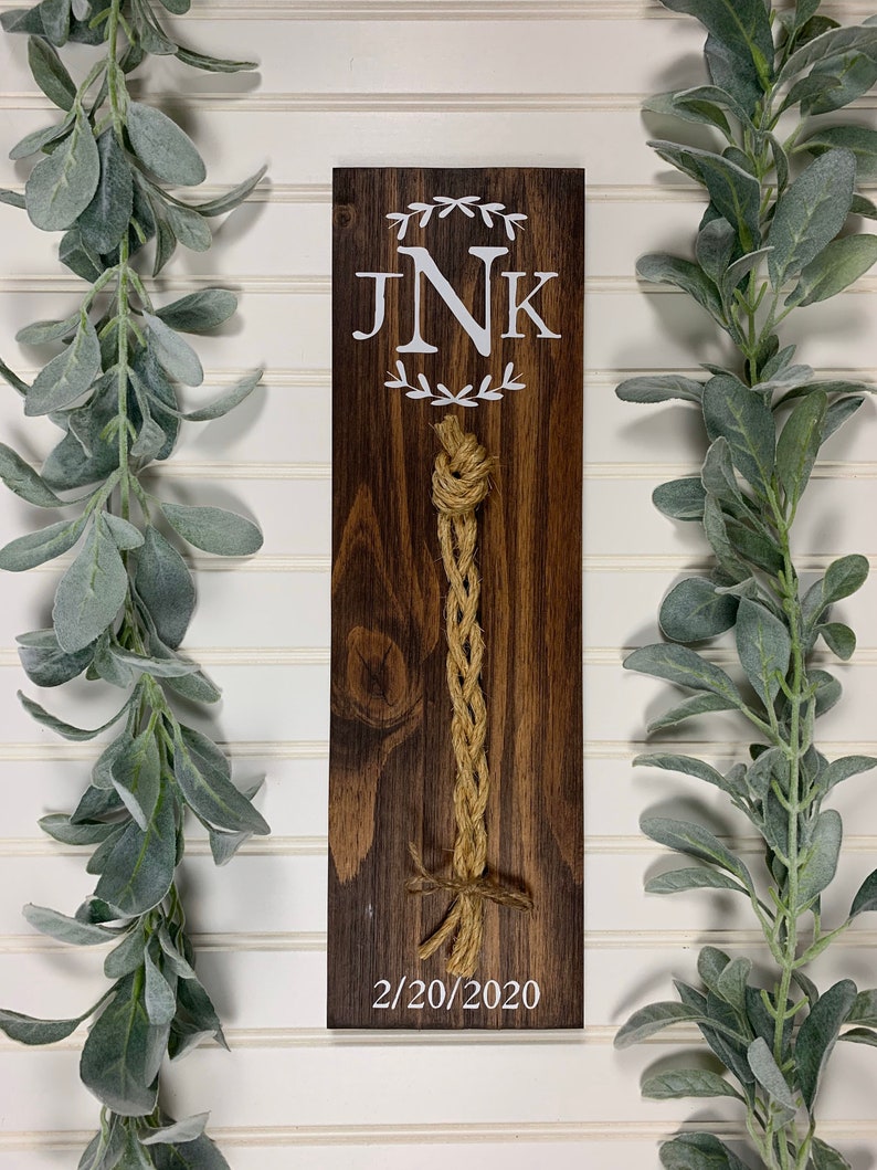 May include: A wooden sign with the initials "JNK" and a date, "2/20/2020", in white paint. A brown rope is tied in a knot and hangs from the sign.