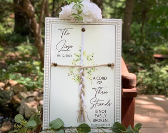 May include: A white wooden sign with a white and brown braided rope tied in a cross shape. The sign has a floral design and the text "The Jays 04/12/2024 A Cord of Three Strands Is Not Easily Broken. Ecclesiastes 4:12"