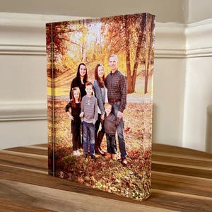May include: A rectangular canvas print featuring a family portrait set against a backdrop of a park with autumn foliage. The family of five is standing in the center, with the parents and three children. The print is displayed on a wooden surface.