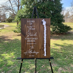 May include: A wooden sign with a white rope braid on the right side. The sign reads "A cord of three strands is not easily broken. Ecclesiastes 4:12" and "The Hiltenbergs EST. 2022".