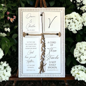 May include: A white framed sign with a rope cross design. The sign has two cards with the names "Dawn & Charlie" and the date "May 18, 2024". The sign also has two cards with the text "So they are no longer two, but one flesh. Therefore what God has joined together. Let no one separate. Matthew 19:6" and "A cord of three strands is not easily broken. Ecclesiastes 4:12".