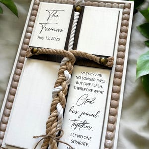 May include: A white wooden frame with a brown wooden cross and a braided rope. The frame has a white background with black text that reads "The Vrainas July 12, 2025" and "SO THEY ARE NO LONGER TWO, BUT ONE FLESH. THEREFORE WHAT God has joined Together. LET NO ONE SEPARATE."