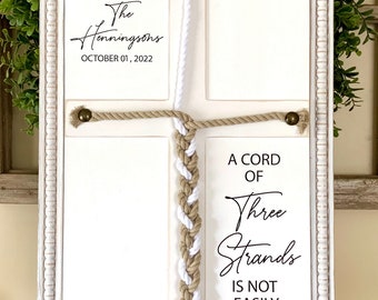 May include: White wooden frame with a white and beige braided rope in the center. The frame is divided into four sections. The top section has the text "The Henningsons OCTOBER 01, 2022". The bottom section has the text "A CORD OF Three Strands IS NOT EASILY BROKEN. ECCLESIASTES 4:12".