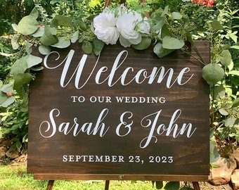May include: A brown wooden wedding sign with white lettering. The sign says "Welcome to our wedding Sarah & John September 23, 2023". The sign is decorated with white flowers and greenery.