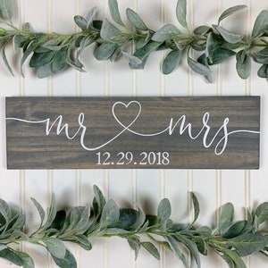 May include: A grey wooden sign with white script reading "mr & mrs", joined by a heart. The date "12.29.2018" is below. The sign is framed by green leafy garlands, suitable for a wedding or anniversary.