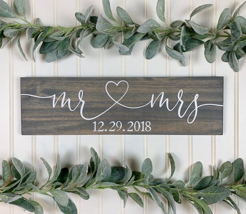 May include: A grey wooden sign with white script reading "mr & mrs", joined by a heart. The date "12.29.2018" is below. The sign is framed by green leafy garlands, suitable for a wedding or anniversary.