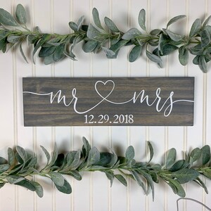 May include: A rectangular wooden sign with the words "mr & mrs" in white script, joined by a heart symbol. The date "12.29.2018" is below. The sign is dark grey and is surrounded by green leafy garlands.