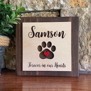 May include: A wooden sign with a brown frame and a light wood center. The sign has the name "Samson" engraved on it, along with a paw print with a red heart in the center. The text "Forever in our Hearts" is also engraved on the sign.