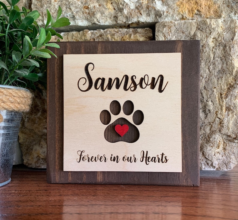 May include: A wooden sign with a brown frame and a light wood center. The sign has the name "Samson" engraved on it, along with a paw print with a red heart in the center. The text "Forever in our Hearts" is also engraved on the sign.