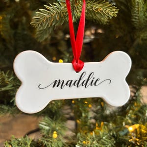 May include: White ceramic dog bone ornament with the name "maddie" written in black cursive script. The ornament is hanging from a red ribbon on a Christmas tree.