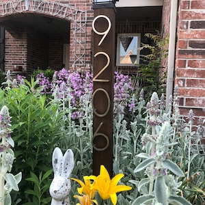 May include: A wooden post with gold metal numbers 9206 attached to it. The post is surrounded by a garden with purple and white flowers, a white rabbit statue, and yellow lilies.