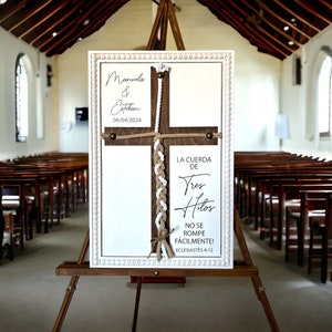 May include: A white sign with a wooden cross and a braided rope. The sign reads "Manuela & Estekan 06/04/2024" and "LA CUERDA DE Tres Hitos NO SE ROMPE F&Aacute;CILMENTE! ECLESIAST&Eacute;S 4:12".