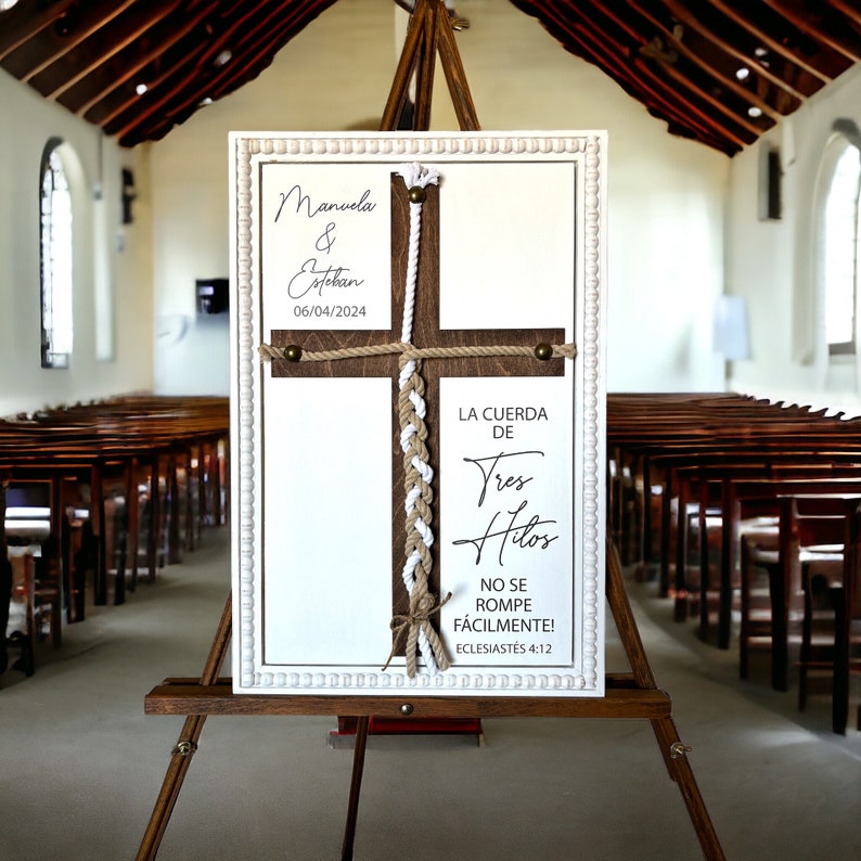 May include: A white sign with a wooden cross and a braided rope. The sign reads "Manuela & Estekan 06/04/2024" and "LA CUERDA DE Tres Hitos NO SE ROMPE F&Aacute;CILMENTE! ECLESIAST&Eacute;S 4:12".