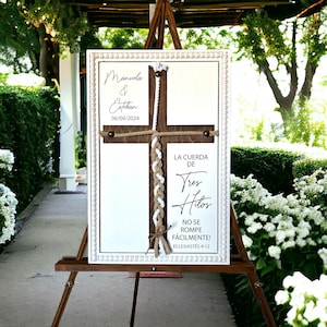 May include: A white wooden sign with a brown wooden cross and a white rope tied in a braid. The sign says "Manuela & Esteban 06/04/2024" and "LA CUERDA DE Tres Hilos NO SE ROMPE F&Aacute;CILMENTE! ECLESIAST&Eacute;S 4:12".