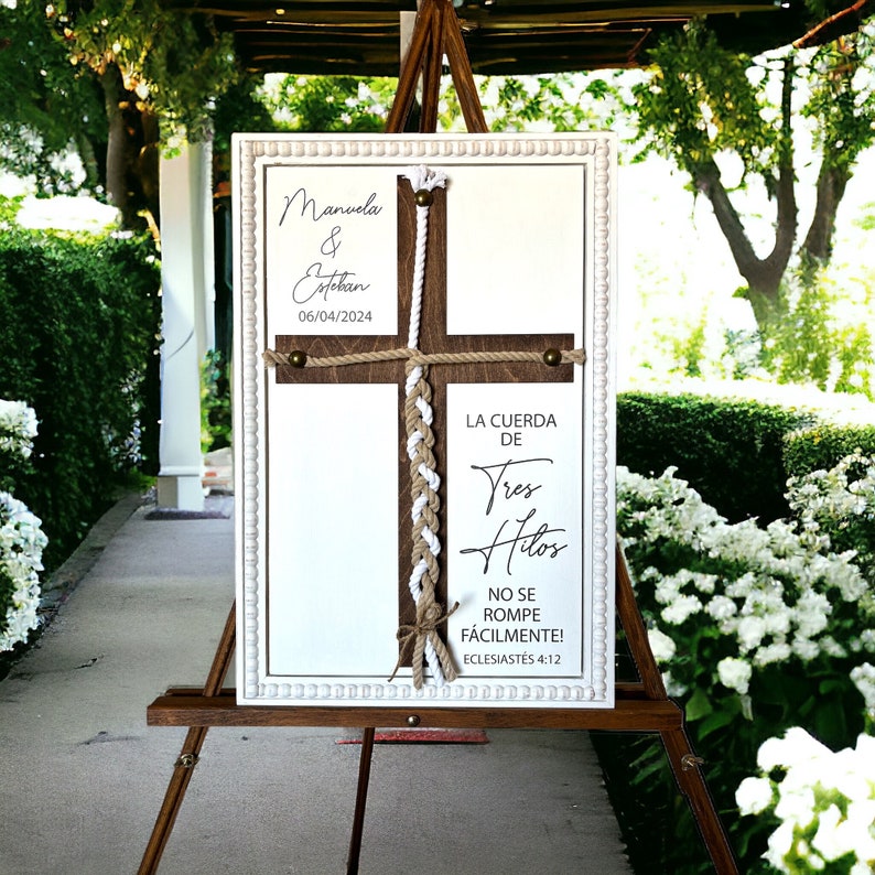 May include: A white wooden sign with a brown wooden cross and a white rope tied in a braid. The sign says "Manuela & Esteban 06/04/2024" and "LA CUERDA DE Tres Hilos NO SE ROMPE F&Aacute;CILMENTE! ECLESIAST&Eacute;S 4:12".
