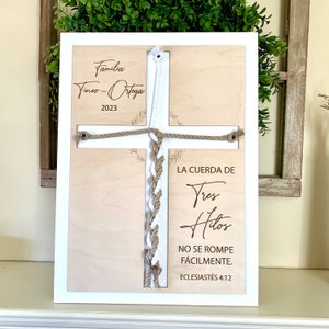 May include: A wooden cross with a white and brown rope tied in a knot. The cross is decorated with the text "Familia Vines-Orteya 2023" and "LA CUERDA DE Tres Hilos NO SE ROMPE FÁCILMENTE. ECLESIASTÉS 4:12".